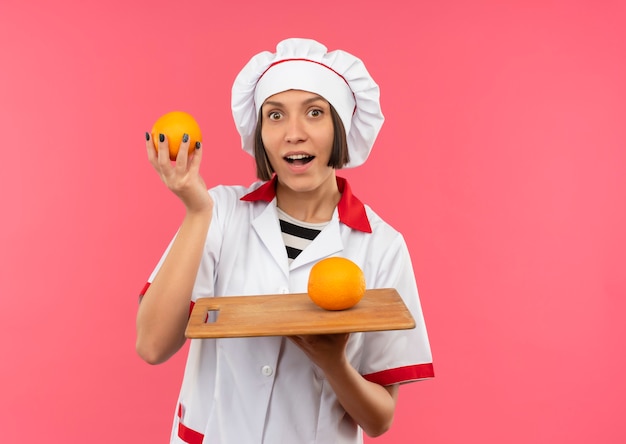 Jovem cozinheira impressionada com uniforme de chef segurando uma laranja e uma tábua com laranja isolada em rosa com espaço de cópia