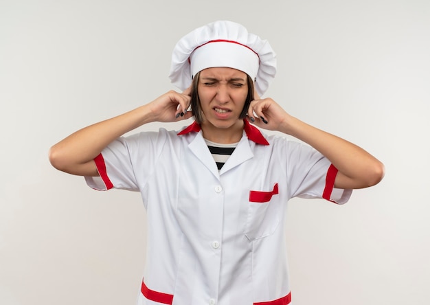 Jovem cozinheira irritada em uniforme de chef, colocando os dedos nas orelhas com os olhos fechados, isolados no branco