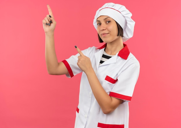 Jovem cozinheira satisfeita com uniforme de chef apontando para cima isolada na parede rosa