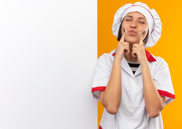Jovem cozinheira satisfeita em uniforme de chef em pé em frente a uma parede branca, colocando os dedos nas bochechas com os olhos fechados, isolados na parede laranja