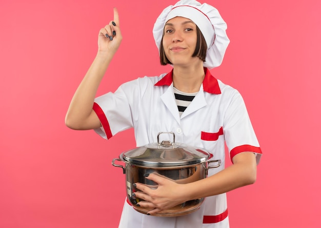Jovem cozinheira satisfeita em uniforme de chef segurando a panela e apontando para cima, isolada na parede rosa