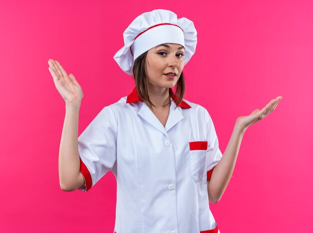 Jovem cozinheira satisfeita usando uniforme de chef, espalhando as mãos isoladas na parede rosa