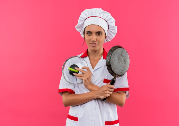 Foto grátis jovem cozinheira satisfeita vestindo uniforme de chef, segurando e cruzando a frigideira com tampa na parede rosa isolada com espaço de cópia