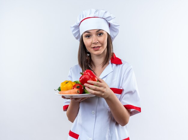 Jovem cozinheira satisfeita vestindo uniforme de chef segurando vegetais no prato isolado na parede branca