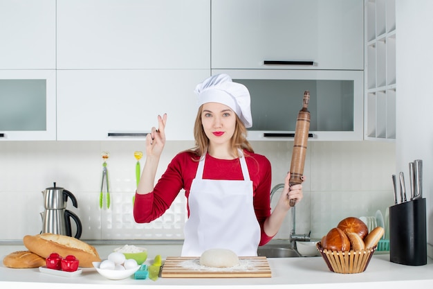 Jovem cozinheira segurando o rolo de massa fazendo sinal de boa sorte na cozinha de frente
