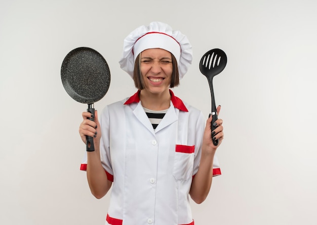 Jovem cozinheira sorridente com uniforme de chef segurando uma espátula e uma frigideira com os olhos fechados, isolada na parede branca