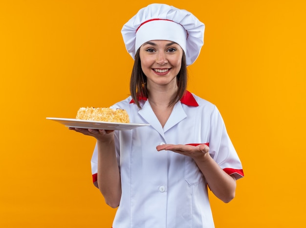 Jovem cozinheira sorridente usando uniforme de chef segurando e aponta com a mão no bolo no prato isolado em fundo laranja