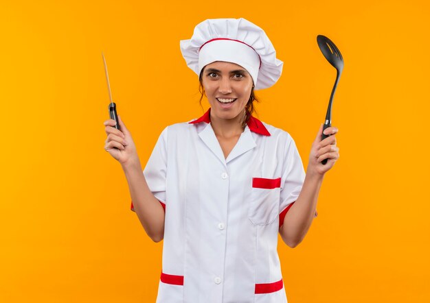 Jovem cozinheira sorridente usando uniforme de chef segurando espátula e faca com espaço de cópia