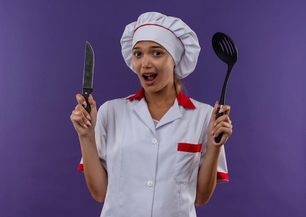 Jovem cozinheira surpresa, vestindo uniforme de chef, segurando uma concha e uma faca em um fundo isolado com espaço de cópia