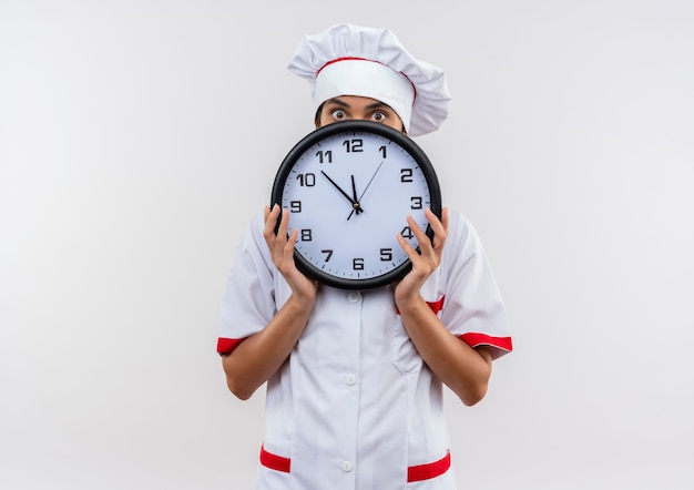 Jovem cozinheira vestindo uniforme de chef coberto com relógio de parede na parede branca isolada com espaço de cópia