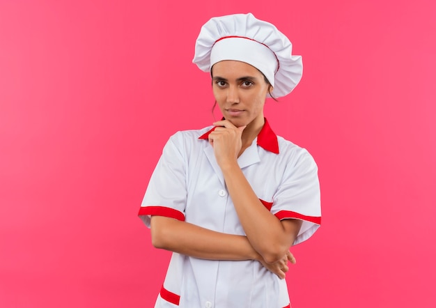 Jovem cozinheira vestindo uniforme de chef, colocando a mão sob o queixo na parede rosa isolada com espaço de cópia