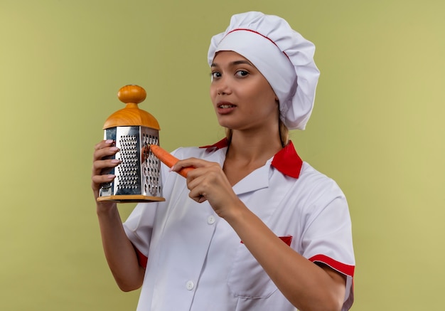 Jovem cozinheira vestindo uniforme de chef ralando cenoura com ralador na parede verde isolada