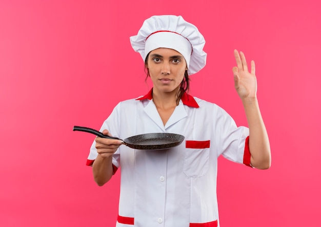 Jovem cozinheira vestindo uniforme de chef, segurando a frigideira e mostrando o gesto ok na parede rosa isolada com espaço de cópia