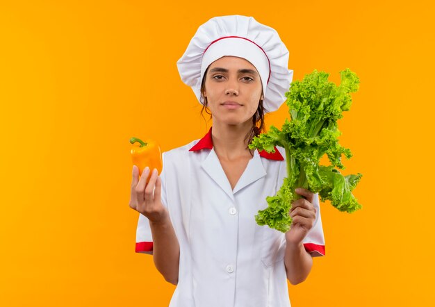 jovem cozinheira vestindo uniforme de chef, segurando salada e pimenta na parede amarela isolada com espaço de cópia