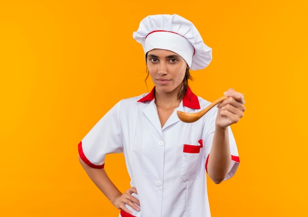 Jovem cozinheira vestindo uniforme de chef segurando uma colher na parede amarela isolada com espaço de cópia