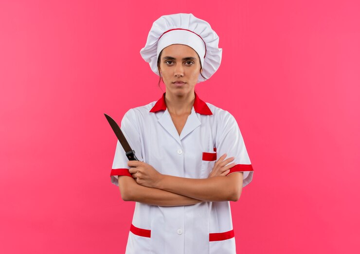 Foto grátis jovem cozinheira vestindo uniforme de chef, segurando uma faca e cruzando as mãos na parede rosa isolada com espaço de cópia