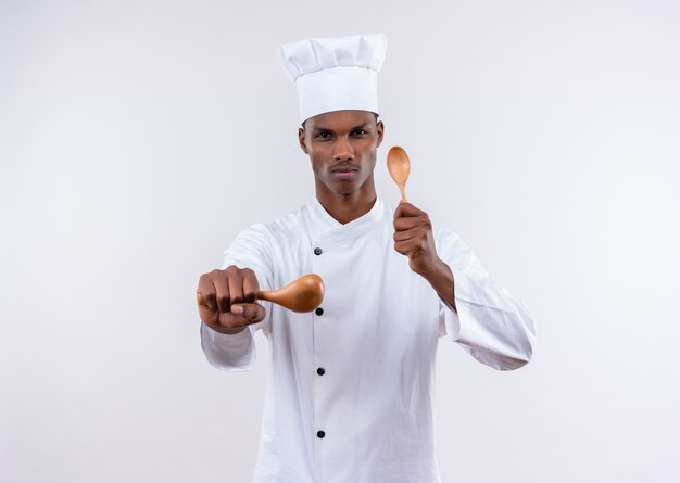 Jovem cozinheiro afro-americano confiante em uniforme de chef finge se defender com colheres de pau em fundo branco isolado com espaço de cópia