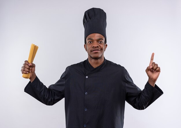 Jovem cozinheiro afro-americano confiante em uniforme de chef segurando um monte de espaguete e aponta isolado no fundo branco com espaço de cópia