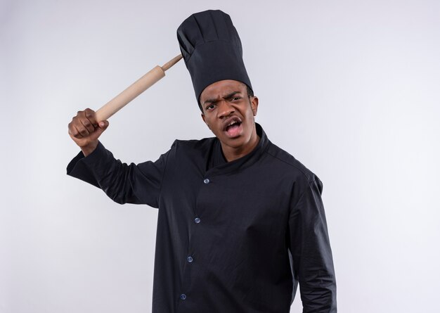 Jovem cozinheiro afro-americano irritado com uniforme de chef segurando o rolo de massa isolado no fundo branco com espaço de cópia