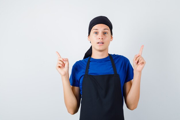 Jovem cozinheiro apontando para cima em t-shirt, avental e olhando desanimado. vista frontal.