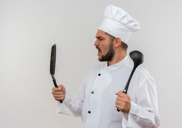 Foto grátis jovem cozinheiro bravo com uniforme de chef segurando uma frigideira e uma concha, olhando para a panela isolada na parede branca