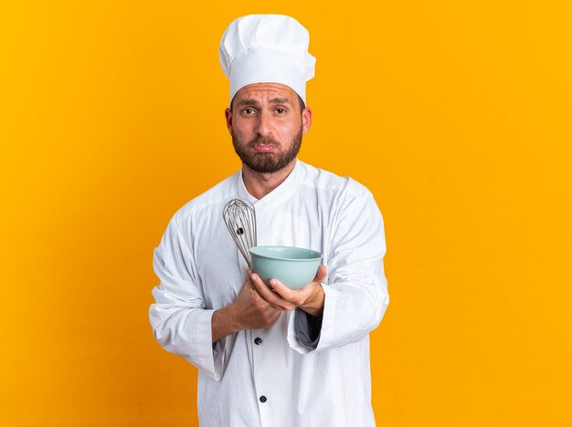 Jovem cozinheiro caucasiano com uniforme de chef e boné segurando um batedor, estendendo a tigela com os lábios contraídos