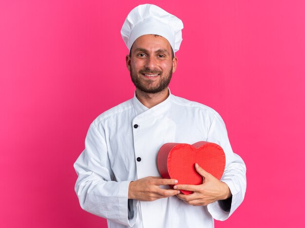 Jovem cozinheiro caucasiano com uniforme de chef e boné segurando um formato de coração a sorrir