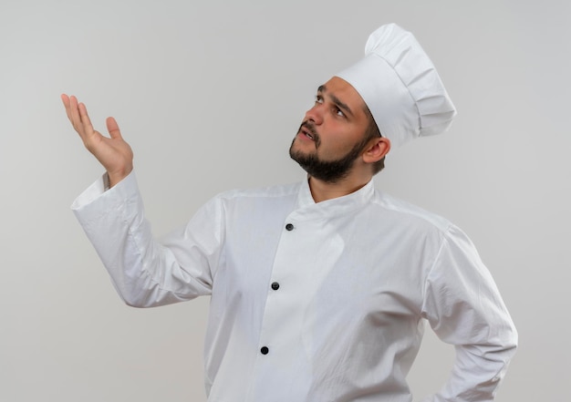 Jovem cozinheiro com uniforme de chef impressionado, olhando para cima e mostrando a mão vazia, isolada na parede branca