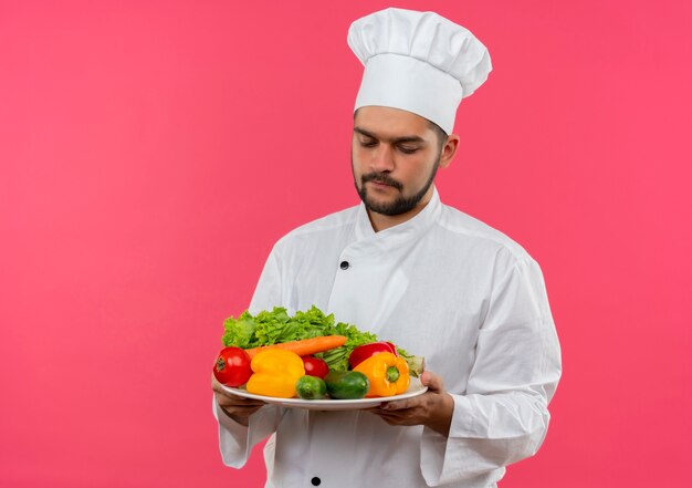 Jovem cozinheiro com uniforme de chef segurando e olhando para o prato de vegetais isolado no espaço rosa