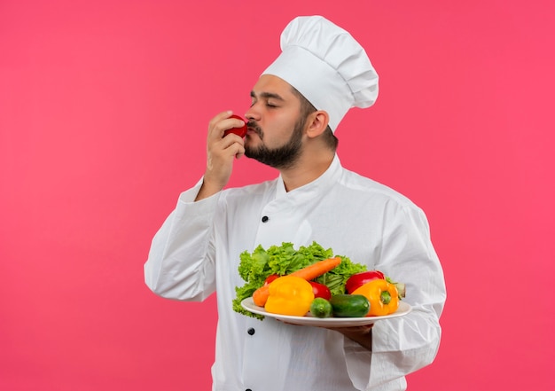 Jovem cozinheiro com uniforme de chef, segurando o prato de legumes e cheirando tomate com os olhos fechados, isolado no espaço rosa