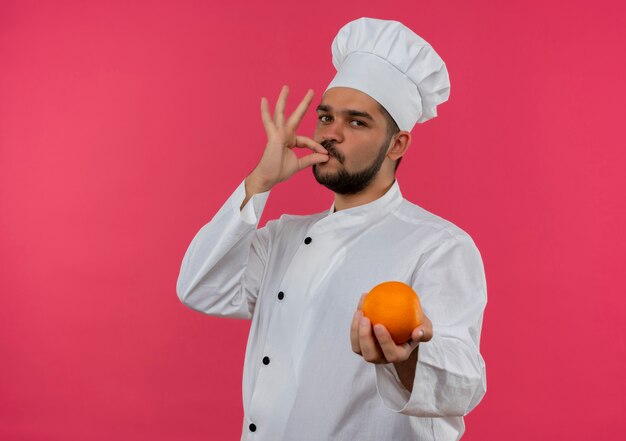 Jovem cozinheiro com uniforme de chef segurando uma laranja e fazendo um gesto saboroso, parecendo isolado no espaço rosa