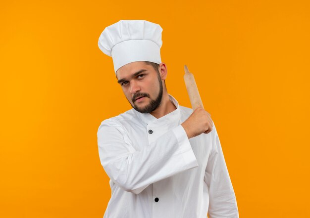 Jovem cozinheiro confiante com uniforme de chef apontando para trás com rolo de massa isolado na parede laranja com espaço de cópia