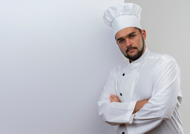 Jovem cozinheiro confiante com uniforme de chef em pé em frente a uma parede branca com postura fechada, parecendo isolado na parede branca com espaço de cópia