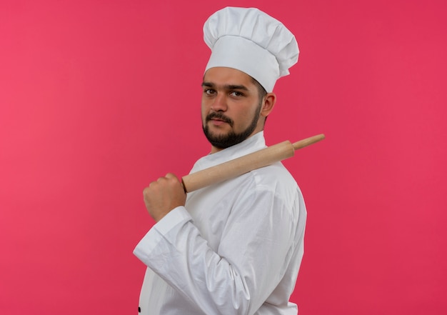 Foto grátis jovem cozinheiro confiante com uniforme de chef em pé na vista de perfil, colocando o rolo no ombro isolado na parede rosa com espaço de cópia