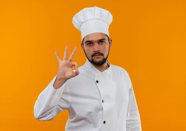 Jovem cozinheiro confiante com uniforme de chef, fazendo sinal de ok e parecendo isolado na parede laranja com espaço de cópia
