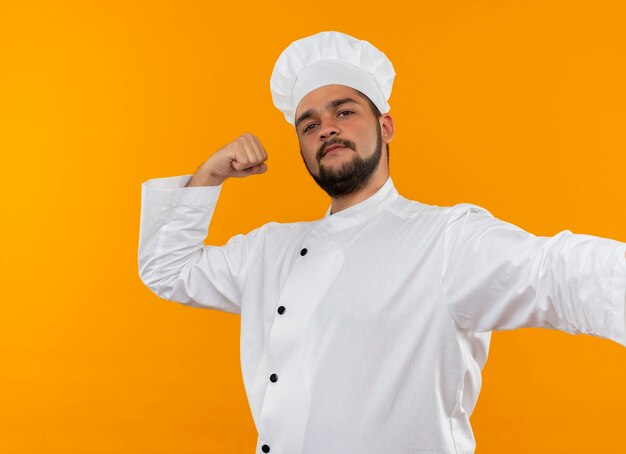 Jovem cozinheiro confiante com uniforme de chef gesticulando forte e isolado na parede laranja