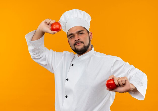 Jovem cozinheiro confiante com uniforme de chef segurando tomates isolados na parede laranja