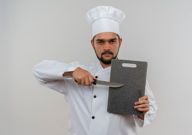 Jovem cozinheiro confiante com uniforme de chef segurando uma tábua de cortar e uma faca isolada na parede branca