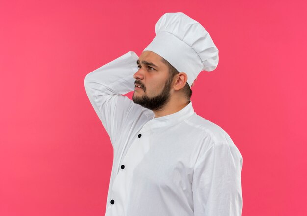 Jovem cozinheiro confuso com uniforme de chef, colocando a mão atrás da cabeça, olhando para o lado isolado na parede rosa com espaço de cópia