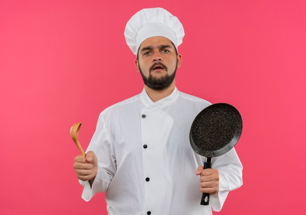 Jovem cozinheiro confuso em uniforme de chef segurando uma frigideira e uma colher isoladas na parede rosa