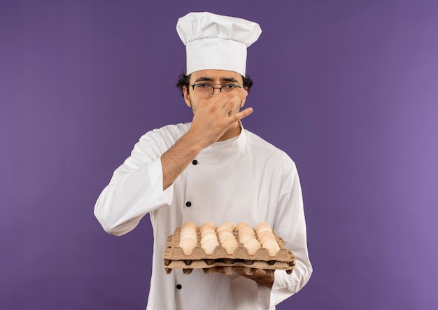 Foto grátis jovem cozinheiro enjoado vestindo uniforme de chef e óculos segurando uma batelada de ovos e nariz fechado na cor roxa