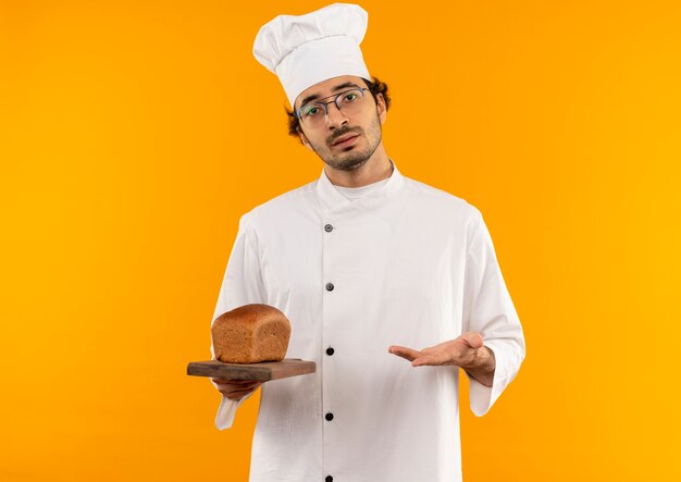 Jovem cozinheiro irritado vestindo uniforme de chef, segurando óculos e apontando com a mão na barba na tábua isolada na parede amarela