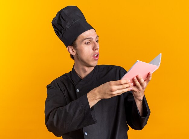 Jovem cozinheiro loiro impressionado com uniforme de chef e boné lendo bloco de notas isolado na parede laranja
