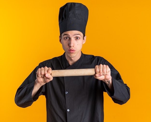 Jovem cozinheiro loiro impressionado com uniforme de chef e boné segurando o rolo de massa horizontalmente, olhando para a câmera isolada na parede laranja