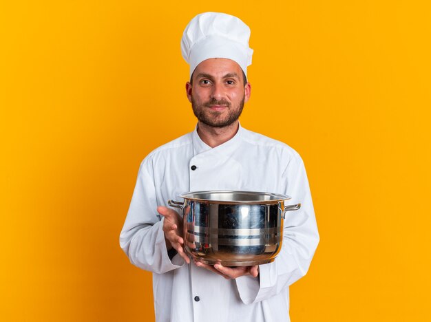 Jovem cozinheiro masculino caucasiano satisfeito com uniforme de chef e boné segurando a panela