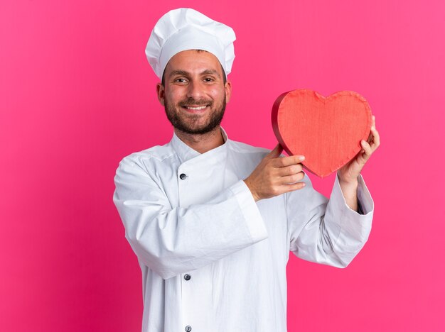 Jovem cozinheiro masculino caucasiano sorridente com uniforme de chef e boné mostrando formato de coração