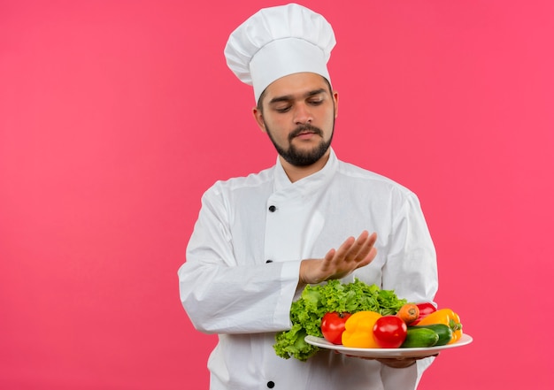 Jovem cozinheiro masculino em uniforme de chef segurando e olhando para o prato de legumes e mantendo a mão acima do prato isolado no espaço rosa