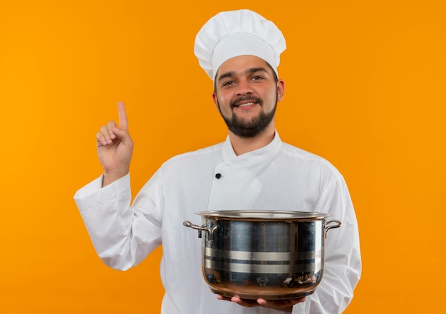 Jovem cozinheiro masculino sorridente com uniforme de chef segurando a panela e olhando para cima, isolado no espaço laranja