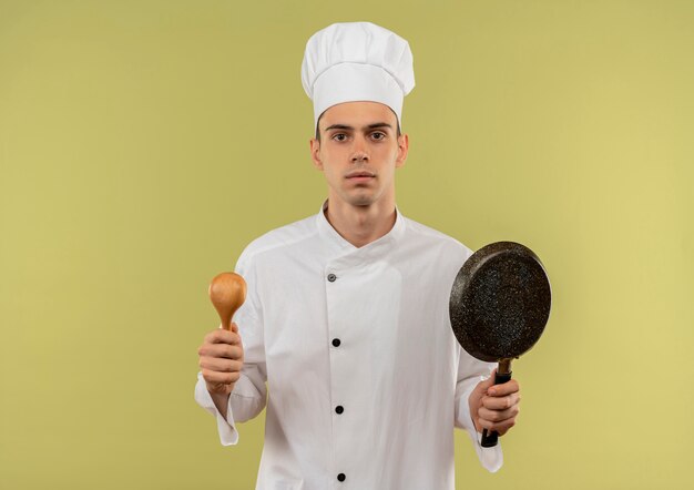 jovem cozinheiro masculino vestindo uniforme de chef, segurando uma concha e uma frigideira na parede verde isolada com espaço de cópia