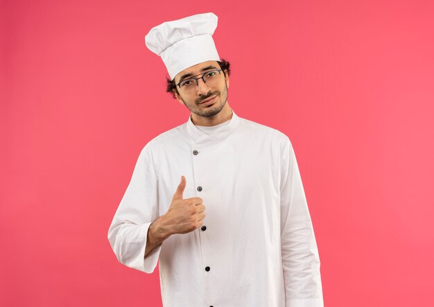 Jovem cozinheiro satisfeito vestindo uniforme de chef e óculos com o polegar isolado na parede rosa